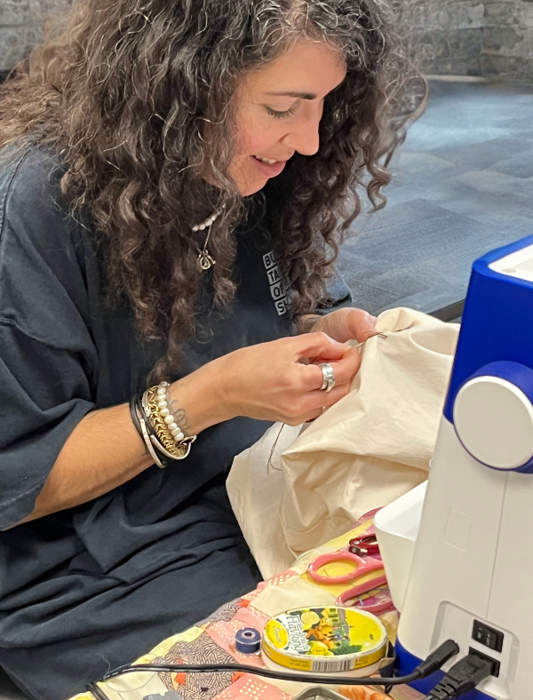 Claudia De Sousa-Baptista of Bushwood Tailors Opportunity Shop in New Bedford is sewing fabric at a table with a sewing machine, scissors, thread, and crafting supplies.